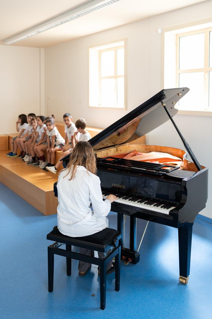 Professora ao piano de cauda, com os alunos sentados em frente